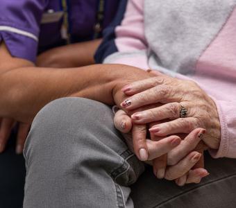 Caregiver in purple uniform gently holding an elderly person's hands.