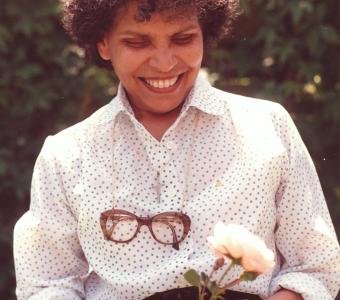 Smiling woman with curly hair holding a light pink rose in a garden.
