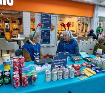 Two women with festive headbands at a market stall selling drinks and snacks.