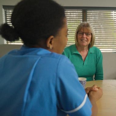 Nurse in blue uniform conversing with two smiling women at a wooden table.
