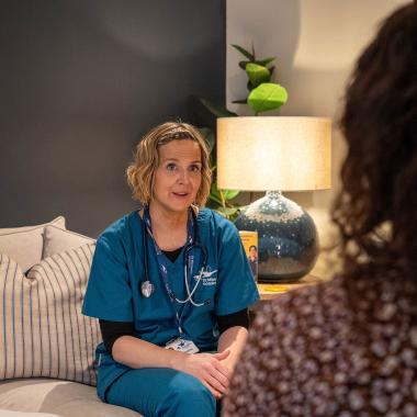 Attentive nurse in teal scrubs with stethoscope sits on a sofa, talking to a patient.