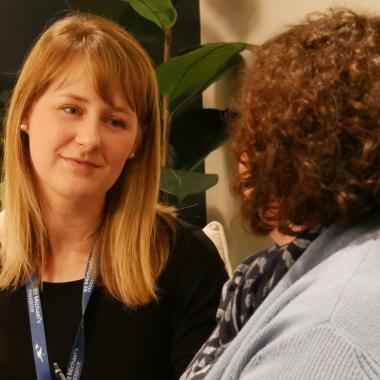 Young woman with blonde hair and lanyard listens to another woman.