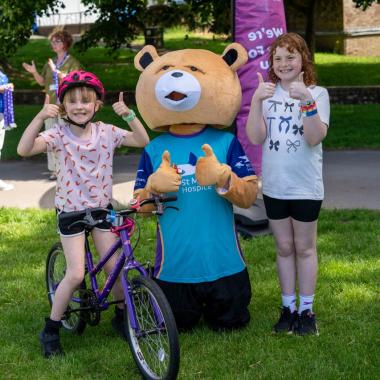 Two girls and a bear mascot give thumbs up with a purple bike on green grass.