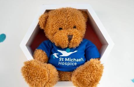 Brown teddy bear in blue "St Michael Hospice" t-shirt, sitting in a white hexagonal shelf.