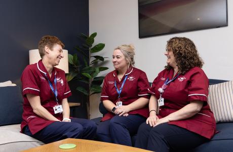 Three smiling women in healthcare uniforms chat on a blue sofa.