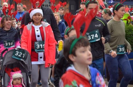 Runners of all ages in festive Christmas attire and race bibs participate in a holiday fun run.