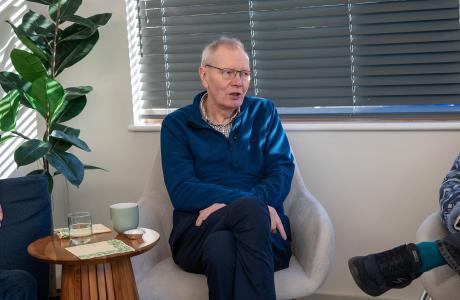 Older man in a blue sweater sits in an armchair, talking in a bright room.