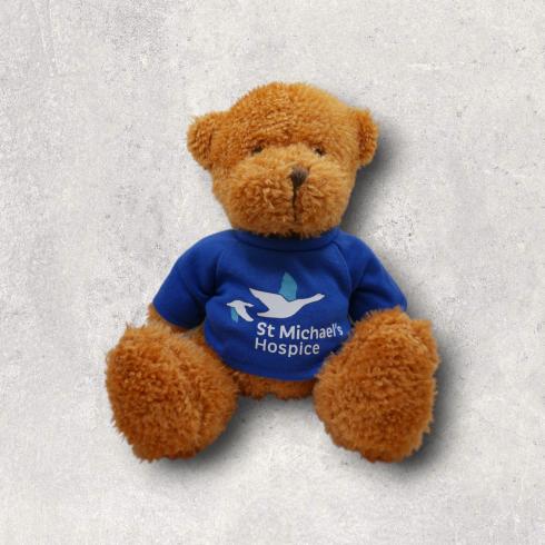 Fluffy brown teddy bear wears blue shirt with St Michael's Hospice logo.