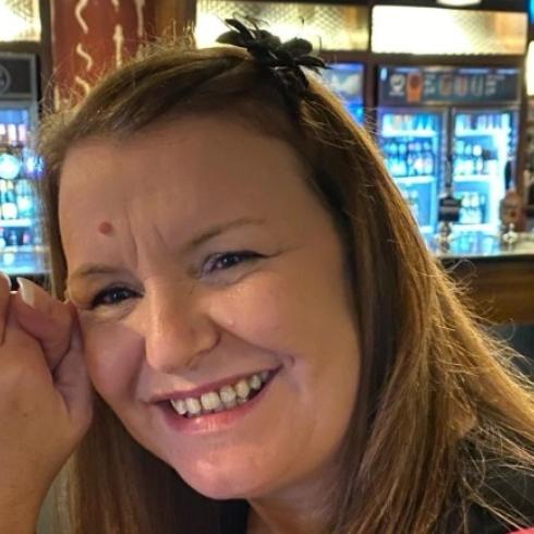 Smiling woman with long hair and clasped hands in a pub, bottles and taps in background.