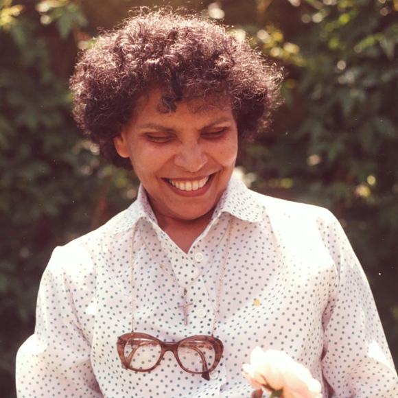 Smiling woman with curly hair holding a light pink rose in a garden.