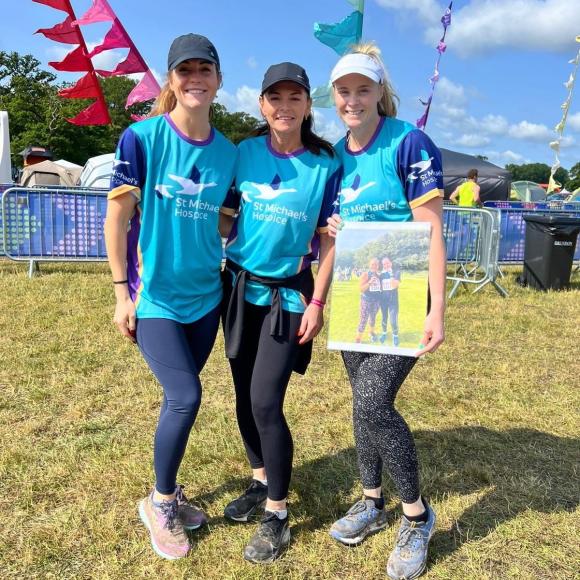 Three smiling women in blue shirts stand on grass, one holds a framed photo. Colorful flags in background.
