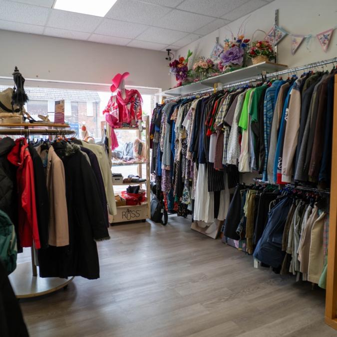 Interior of a small clothing store with racks of various garments and a bright window.