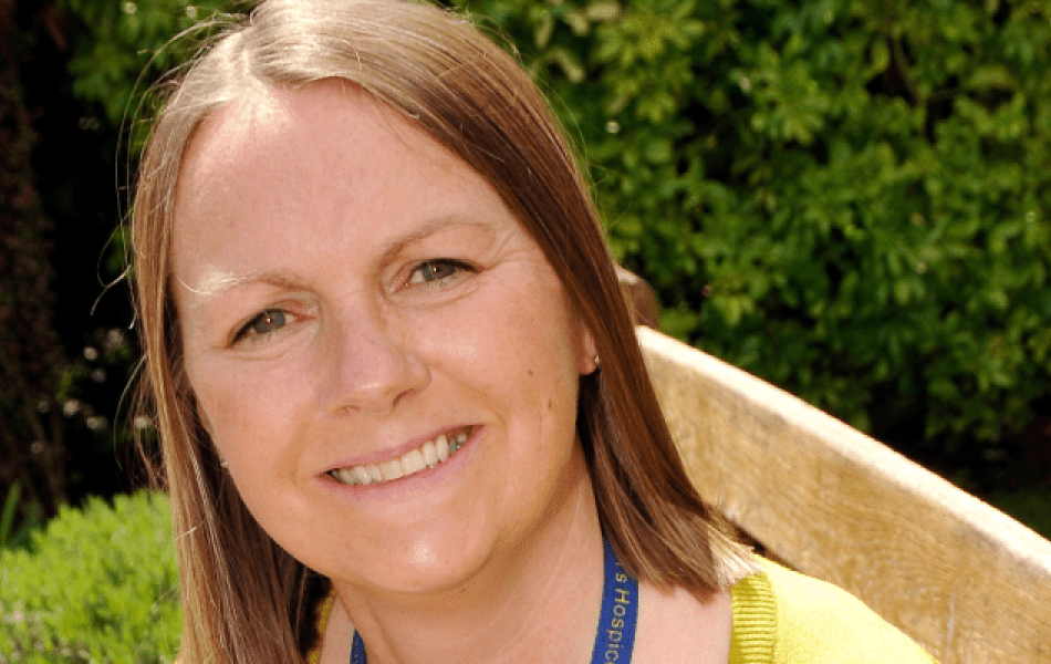 Smiling woman with long brown hair, yellow top, and blue lanyard outdoors.