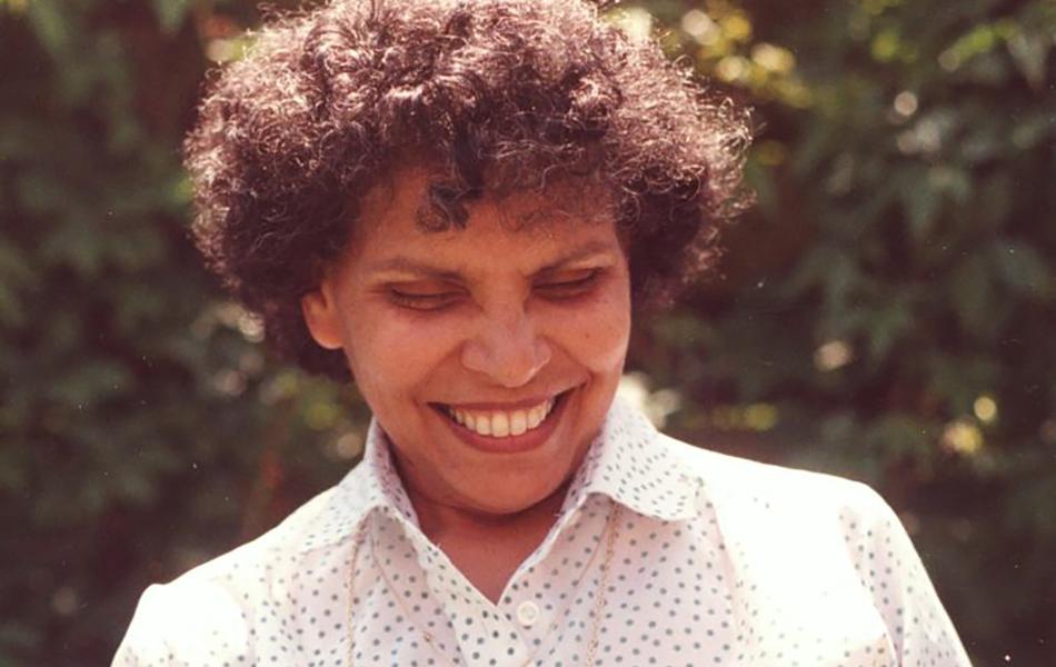 Smiling woman with curly hair holding a light pink rose in a garden.