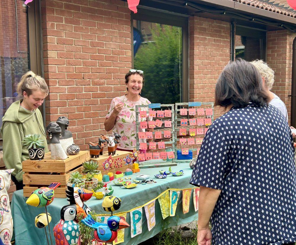 Woman smiles behind a colorful craft stall with handmade birds and plants.