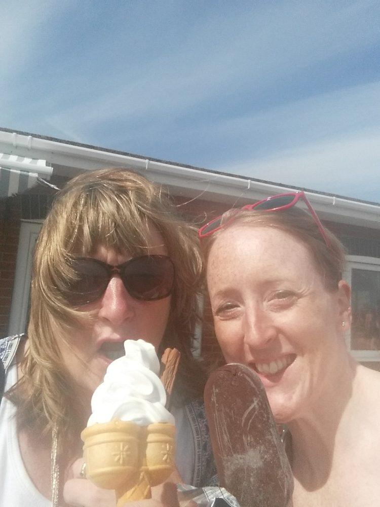 Two happy women eating ice cream, one with a cone, one with a bar, on a sunny day.