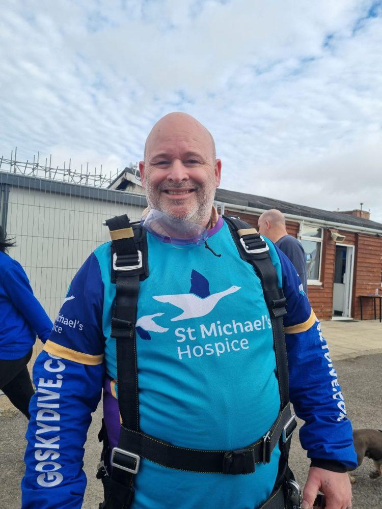 Smiling man in blue and teal skydiving suit for St Michael's Hospice, ready for a jump.