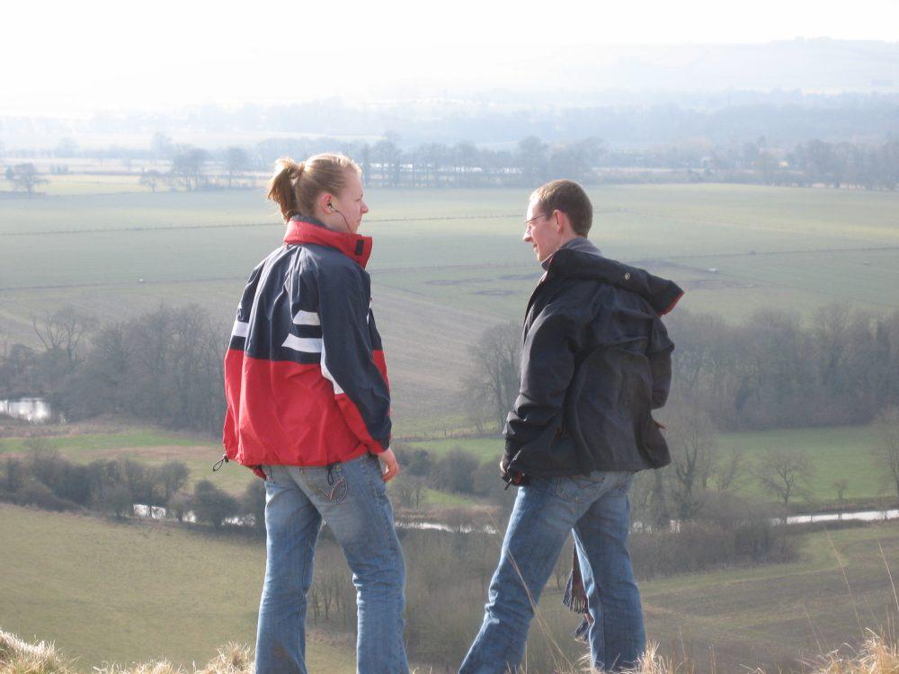 Two people laugh on a hill overlooking a green valley.