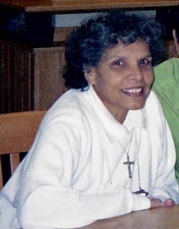 Smiling woman with dark curly hair wears a white top and cross necklace at a wooden table.