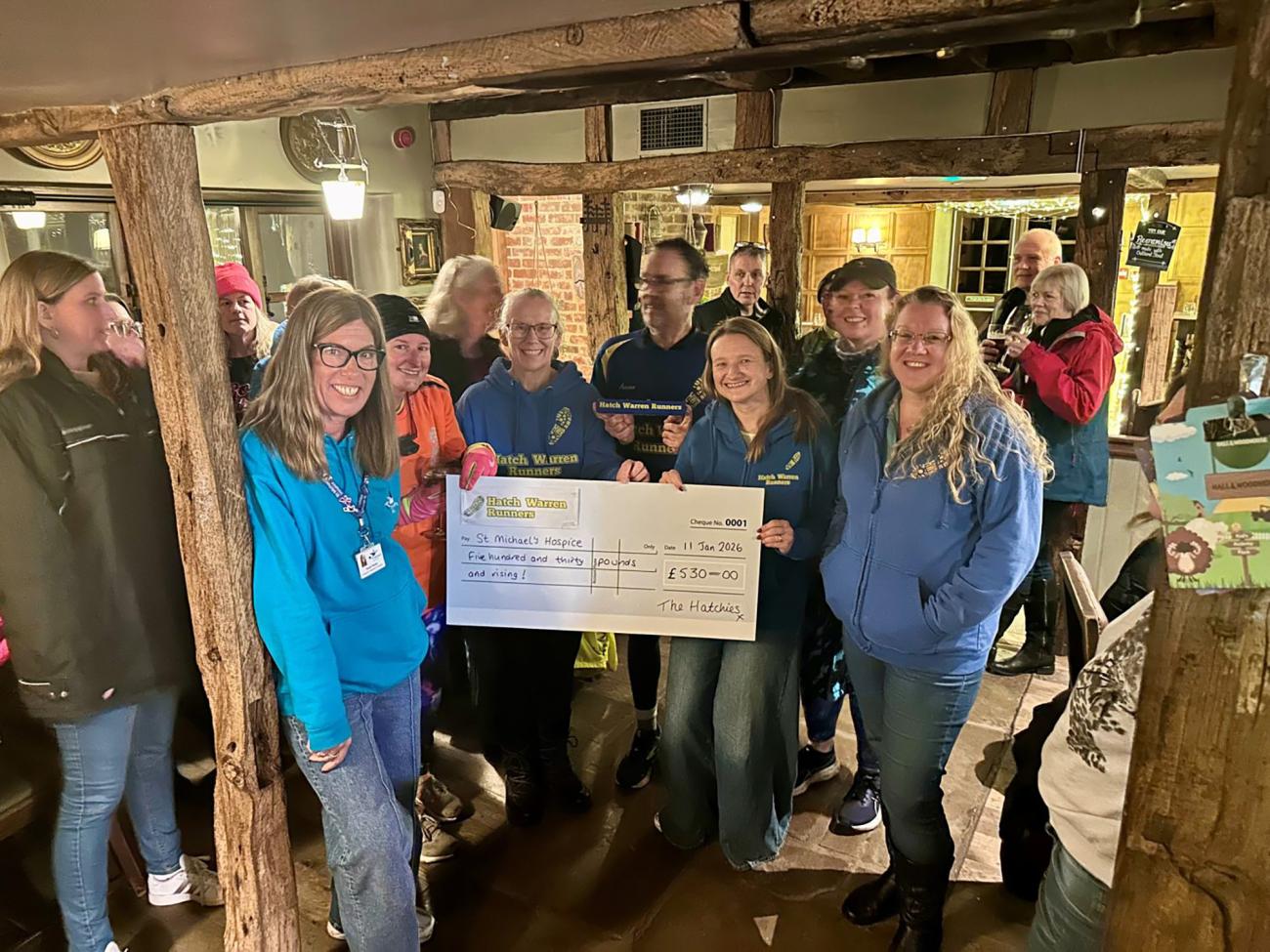 Group of smiling people, two women holding a large novelty check in a rustic pub.