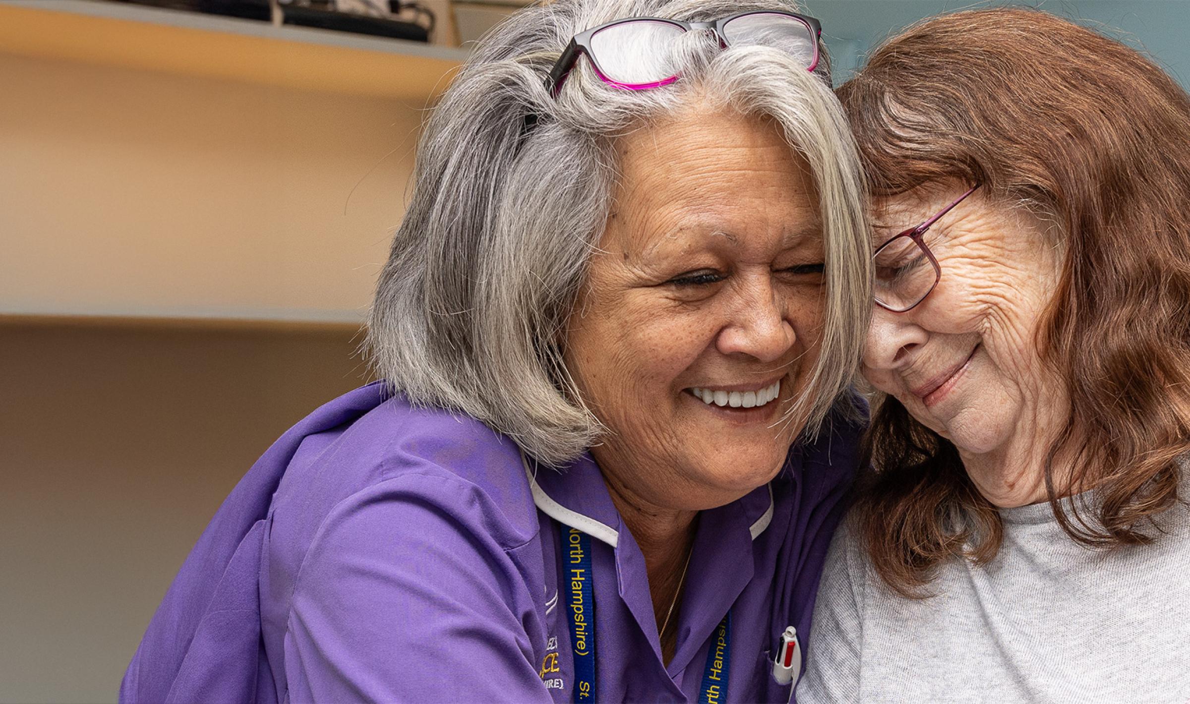 Happy caregiver in purple embraces older woman, both smiling warmly.