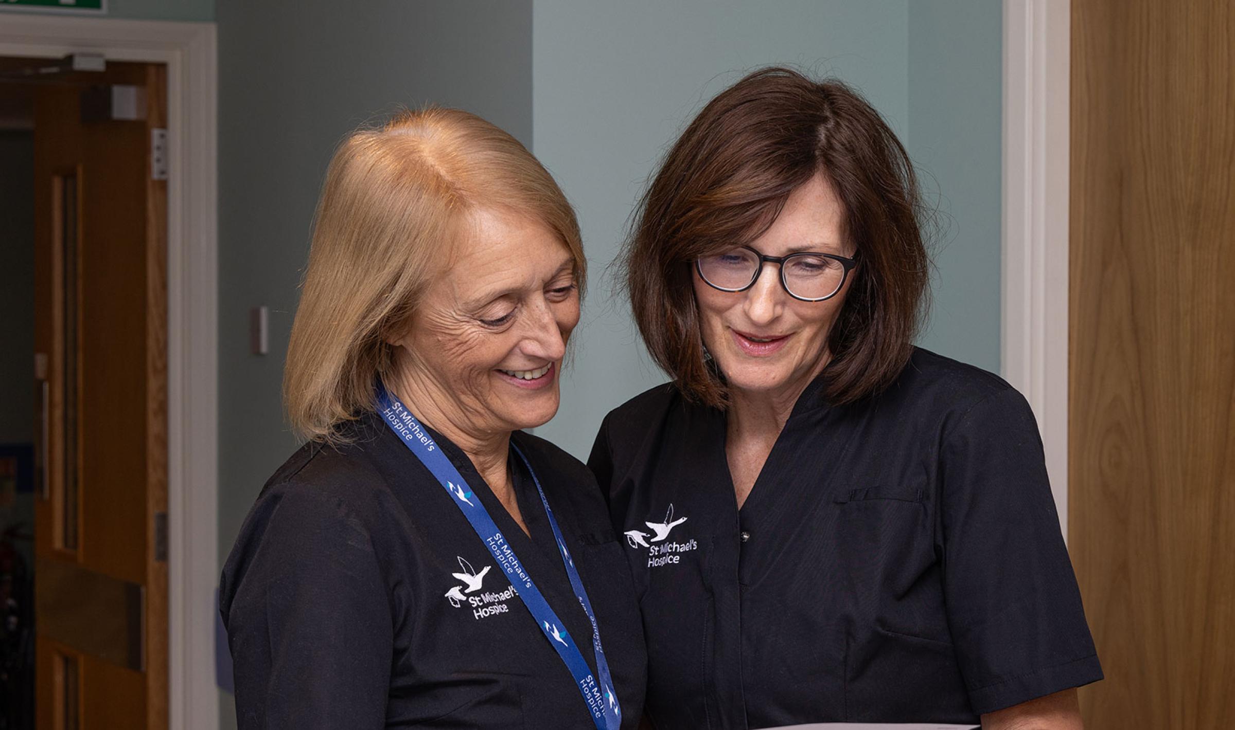 Two smiling women in dark uniforms reviewing a document in a hallway.