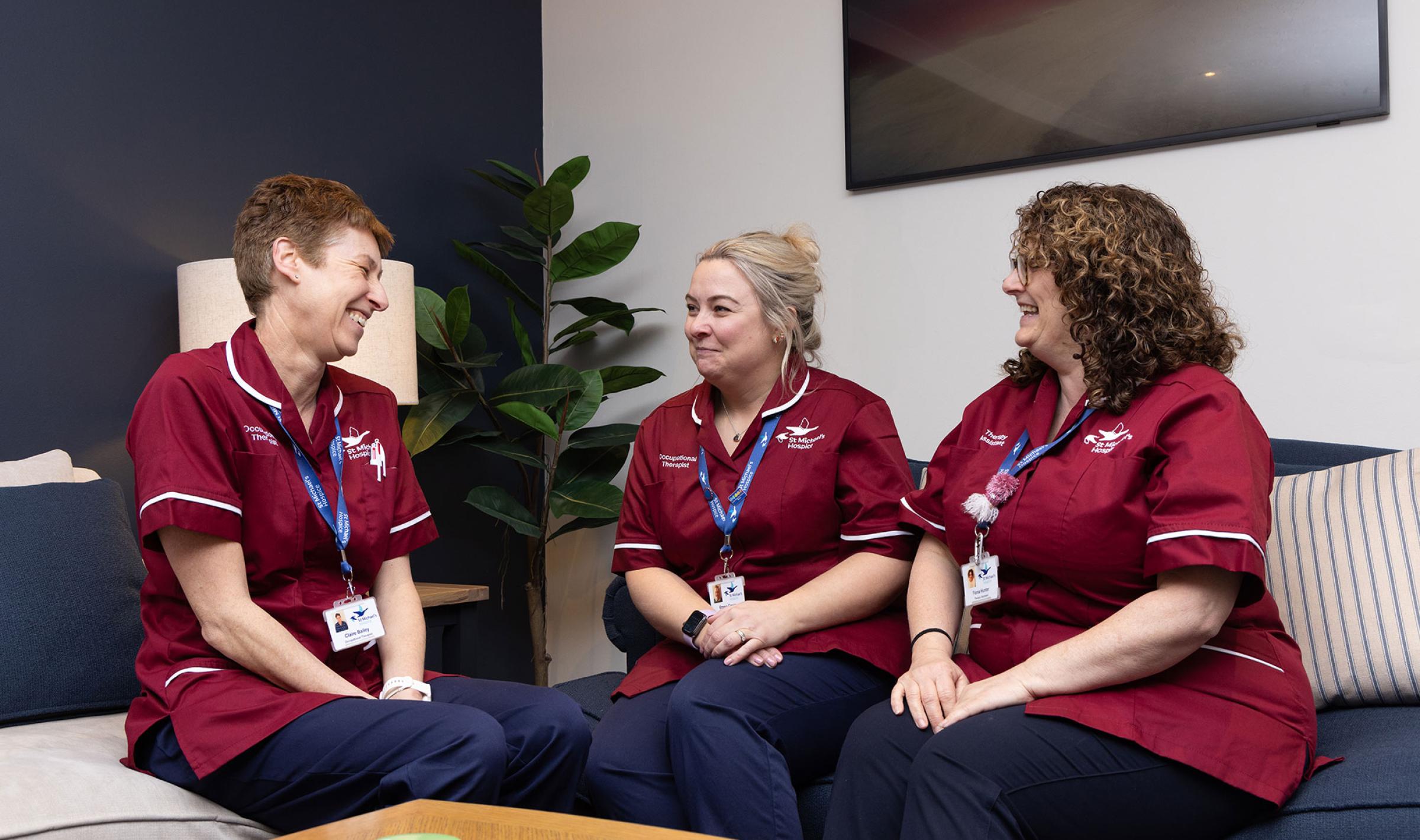 Three smiling nurses in maroon scrubs chat on a blue sofa in a lounge.