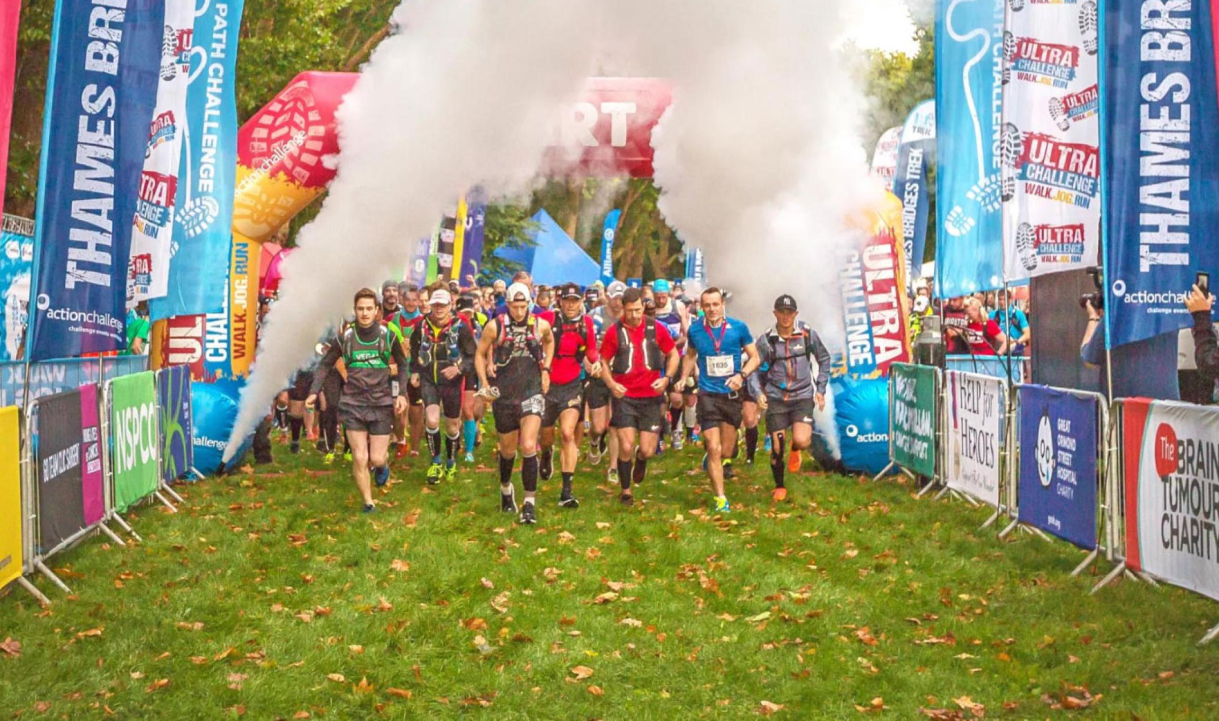 Runners burst through white smoke at a race start line lined with colorful event banners.